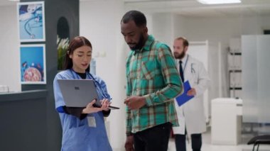 Asian nurse and african american patient talking about disease diagnosis and looking at laptop to find treatment support. Medical assistant consulting man to help with medication. Handheld shot.