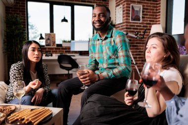 En iyi arkadaşlarla iş terfisini kutlarken kanepede oturan gülümseyen Afrikalı Amerikalı adam. Arkadaşlık yıldönümümüzü kutlamak için bir grup insan evde şarap partisi veriyor..