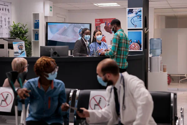 Young man at hospital reception talking to medical staff, discussing about checkup appointment with nurse and receptionist. Male patient in waiting room area during covid 19 pandemic.