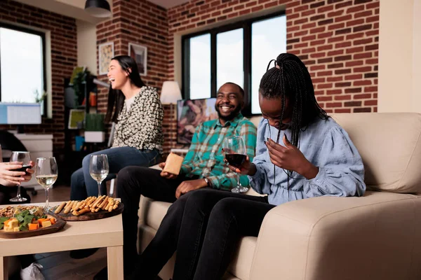 Cheerful diverse people laughing together at wine party in living room at home. Multiethnic group of friends smiling heartily while celebrating birthday event with alcoholic beverages and snacks.