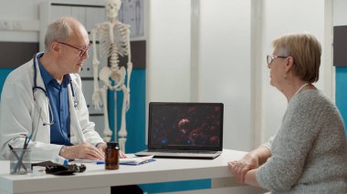 Genel pratisyen, dizüstü bilgisayarda Coronavirus Illustrasyonunu açıklıyor, kabinedeki emekli kadınla konuşuyor. Muayene randevusunda Covid 19 virüs animasyonu gösterimde.