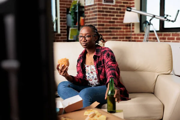 Young person eating burger meal and drinking bottle of beer in front of tv, watching movie or film on channel program. Eating fast food from takeaway delivery package, having fun.