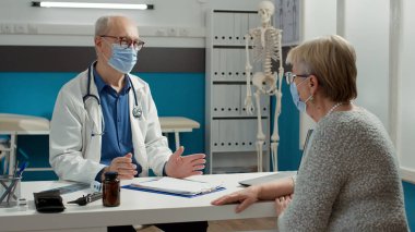 Erkek doktor, yaşlı bir kadınla check-up yapıyor. Hastalık teşhisi ve reçeteli tedavi hakkında konuşuyor. Doktor muayene randevusunda hastaya sağlık desteği veriyor.