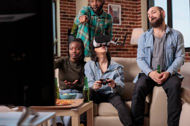 Diverse group of friends playing video games with vr glasses, using augmented reality and 3d simulation to play competition. Cheerful people enjoying headset and game at house party.