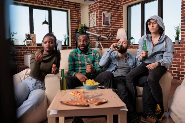 Diverse group of sport fans watching football match game, drinking bottles of beer and cheering to win soccer championship. Friends enjoying pitch tournament competition on television.