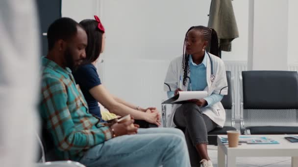 アフリカ系アメリカ人の専門家は 待機地域のロビーで母親と少女に相談するための医療に関する質問をする 医者は病気や診断について母親と子供と話す — ストック動画