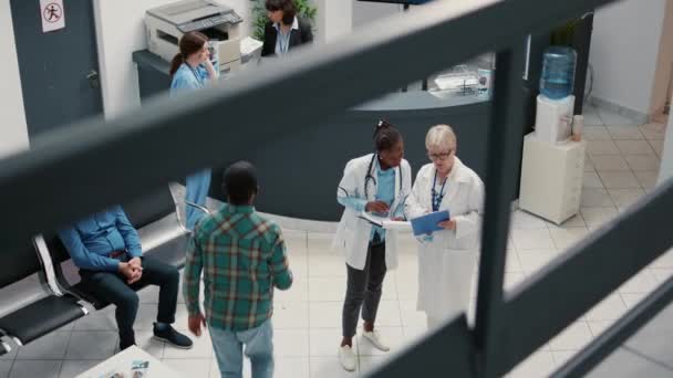 病院の受付ロビーで高齢の医師とアフリカ系アメリカ人の医療従事者が話し合い 病気の患者と相談するための医療報告書をチェックする 待合室の専門知識を持つ医療スタッフ — ストック動画
