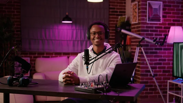 Portrait of african american online radio host sitting at desk in home recording studio with boom arm microphone and digital audio mixer. Podcaster smiling confident at camera while broadcasting live.