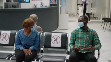 African american patients with face mask talking in waiting area, preparimg to start checkup examination with medic during covid 19 pandemic. Man and woman sitting in hospital reception.