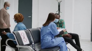 Çok ırklı maskeli bir grup insan hastane resepsiyonunda oturmuş koronavirüs salgını sırasında randevuya katılmak için bekliyorlar. Bekleme alanındaki hastalar covid 19 önleme hakkında konuşmak için bekliyor..