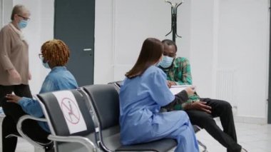 Multiethnic group of people with face masks sitting in hospital reception, waiting to attend appointment during coronavirus pandemic. Patients in waiting area to discuss about covid 19 prevention.