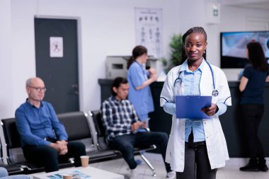 Afro-Amerikan tıp doktorunun portresi laboratuvar önlüğüyle poz veriyor ve elinde bir sonraki hasta randevusunu bekliyor. Steteskoplu doktor yoğun bir hastane resepsiyonunda bekliyor..