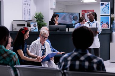 Hastanenin resepsiyonunda rutin check-up randevusu için anne ve çocuk formunu dolduran dosya doktoruyla birlikte kıdemli çocuk doktoru. Modern özel klinikte bekleyen farklı insanlar var..