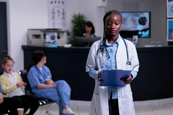 Portrait of african american doctor with stethoscope looking at clipboard with patient data in hospital reception. Confident medic in clinic reception lobby waiting for patient appointment.