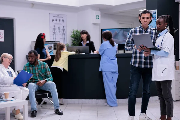 Medic holding laptop with lab results talking with asian patient while older doctor is admitting patient in private clinic. Medical specialist having conversation with young man checking appointment.