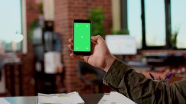 Businessman holding greenscreen display on mobile phone, analyzing blank mockup template with isolated copyspace. Modern smartphone with chroma key background screen used in startup office.
