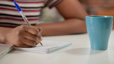 Female freelancer writing lesson information on notebook at desk, taking notes to learn about startup business online on school network. Woman using textbook. Handheld shot. Close up.