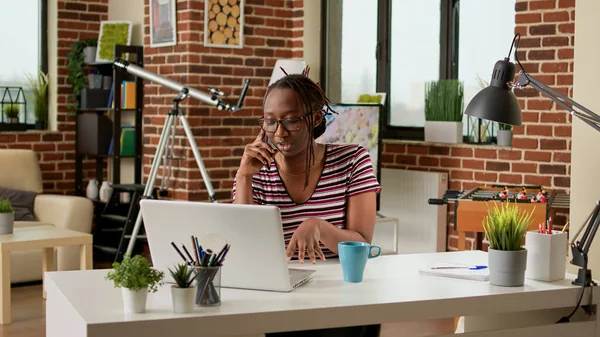 Female freelancer answering phone call on smartphone, working remotely on laptop from home. Businesswoman doing remote telecommunications job and talking on telephone line at desk.