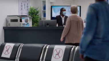 African american receptionist with face mask working on appointments to help with healthcare service and support. Hospital employee analyzing checkup reports at reception desk.