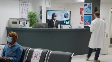 Diverse group of patients with face masks sitting in hospital reception, talking to receptionist about medical appointment. People doing consultation during coronavirus epidemic.