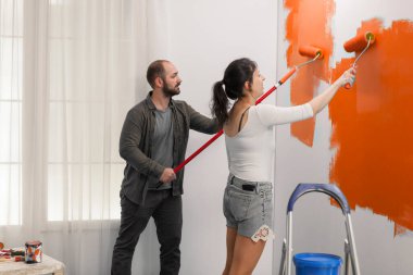 Young couple using orange color to paint wall with roller and paintbrush during home renovation. Painting with new color to redecorate apartment room, interior renovation and house improvement.