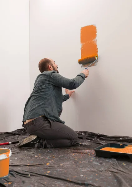 Man painting wall with orange paint, using paintbrush and roller to redecorate apartment room. Renovating home with diy tools and equipment. Vibrant paint renovation and improvement.