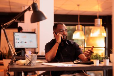 Afrikalı Amerikalı işadamı cep telefonuyla konuşuyor, ofisteki müdürle uzaktan konuşuyor. Akıllı telefondan aranan, kağıt işleri hakkında konuşmak için telefon hattını kullanan.