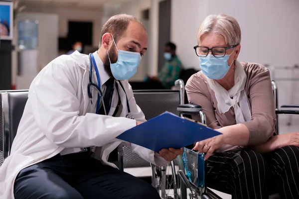 Physician talking to senior patient with chronic disability, consulting old woman suffering from impairment. Female wheelchair user having medical appointment with medic during covid 19 epidemic.