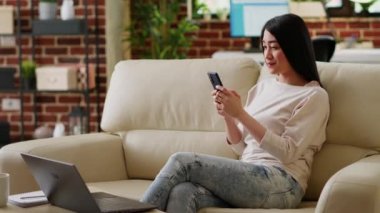 Uzaktan kumandalı telefon kullanan ve evdeki koltukta oturan güzel bir kadın. Dokunmatik ekran modern cihazlı çekici kişi oturma odasındaki kanepede dinlenirken uzaktan çalışmanın tadını çıkarıyor..