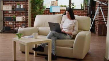 Focused young adult asian student attending online class while typing important lessons on modern portable computer at home. Beautiful woman sitting on sofa while working remotely on laptop.