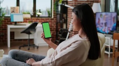 Kanepede otururken yeşil ekranlı akıllı telefonu olan mutlu, güler yüzlü bir kadın. Cep telefonuyla koltukta oturan neşeli kişinin arka planı sahte kroma..