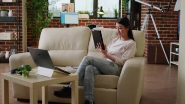Happy asian woman having modern tablet browsing internet while sitting on couch at home. Cheerful smiling heartily person with touchscreen device surfing webpages while doing remote work.