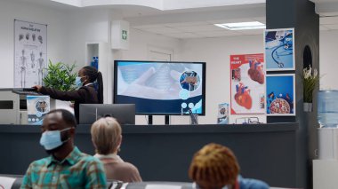 Yüzü maskeli Afrikalı Amerikalı resepsiyonist, sağlık hizmeti ve desteğe yardımcı olmak için randevular üzerinde çalışıyor. Hastane çalışanı resepsiyondaki sağlık raporlarını inceliyor..