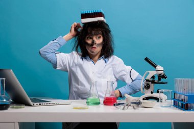 Kafasında cam test tüpleri olan tepsiyle aptal gibi davranan komik görünümlü kimyagerin portresi. Laboratuvar patlamasından sonra masasında oturan aptal suratlı kaçık bilim adamı..