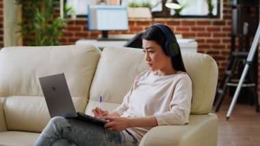 Focused asian woman taking notes while working remotely and listening to music at home in living room. Attentive person doing remote work on laptop while writing down important job informations.