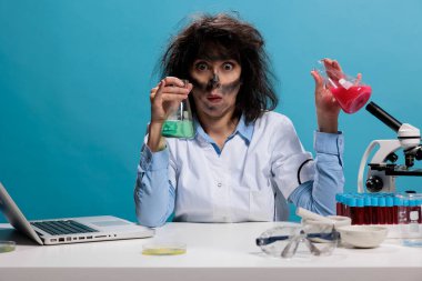 Deneysel sıvı bileşiklerle dolu cam kavanozlar varken masasında oturan aptal görünümlü çılgın bir laboratuar çalışanı. Komik yüz ifadesi olan aptal bilim adamı serumla dolu şişeleri tutuyor..