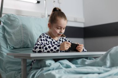 Akıllı telefondan oyun oynarken yatakta dinlenen güzel hasta kız. Pediatrik sağlık kliniği koğuşunda oturup çizgi film izleyen hasta çocuğu hastaneye kaldırdık..