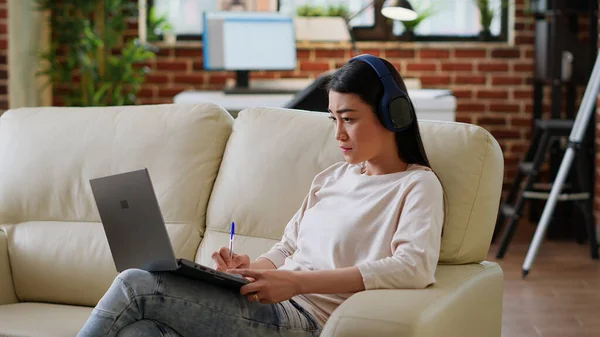 Focused Asian Woman Taking Notes While Working Remotely Listening Music — Fotografia de Stock