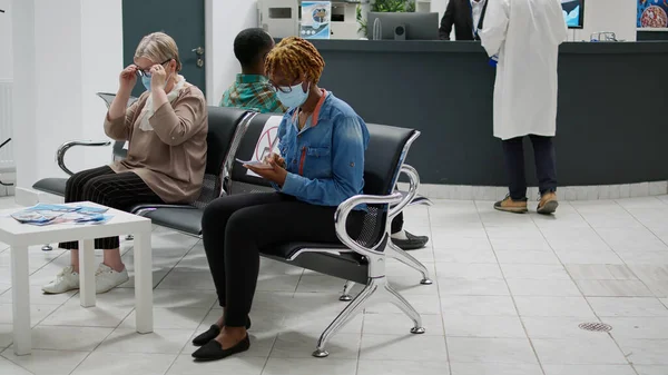 Diverse group of patients with face masks sitting in waiting area at hospital reception, preparing to start medical checkup appointment during covid 19 pandemic. Healthcare exam.
