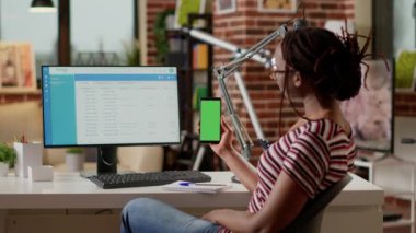 Genç bir çalışan cep telefonundan yeşil ekran ekranına bakıyor, masasında uzaktan kumanda yapıyor. İzole edilmiş model şablonu, boş krom anahtar görüntüsü ve telif alanı arkaplanı ile çalışıyor. Üçayak atışı.