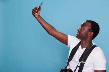Afro-Amerikan profesyonel fotoğrafçı mavi arka planda dururken akıllı telefon cihazıyla selfie çekiyor. DSLR kamerasına sahip kendine güvenen bir insan telefonuyla kendi fotoğrafını çekiyor..