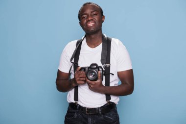 Kendine güvenen fotoğraf makinesi olan neşeli, yakışıklı, Afrikalı etnik kökenli adam. Kendine güvenen profesyonel fotoğrafçı modern kameraya sahipken içten bir gülümsemeyle mavi arka planda duruyor..