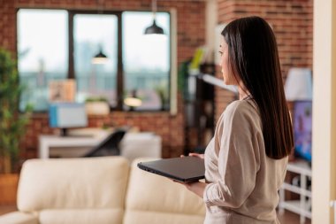 Otantik güzel Asyalı kadın elinde dizüstü bilgisayarla oturma odasında yürüyor uzaktan çalışmaya başlıyor, internet kullanıcısından çalışıyor. Merkez ofisteki serbest çalışan biri işe başlıyor.