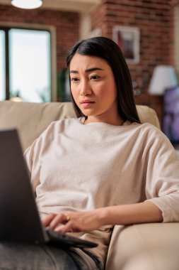 Asyalı etnik kökenli bir kadın ev ofisinde dizüstü bilgisayar teknolojisi kullanarak internet üzerinden internet üzerinden sosyal medyada dedikodu okuyarak çalışıyor.