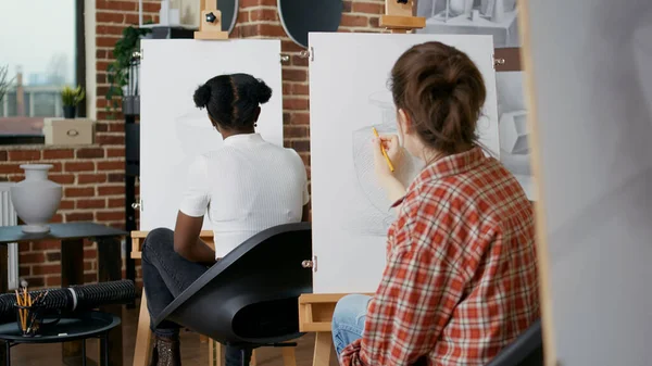 Young woman using pencil to draw vase on white canvas, learning new artistic skills in art class. Student drawing inspiration model at lesson practice with multi ethnic group of people.