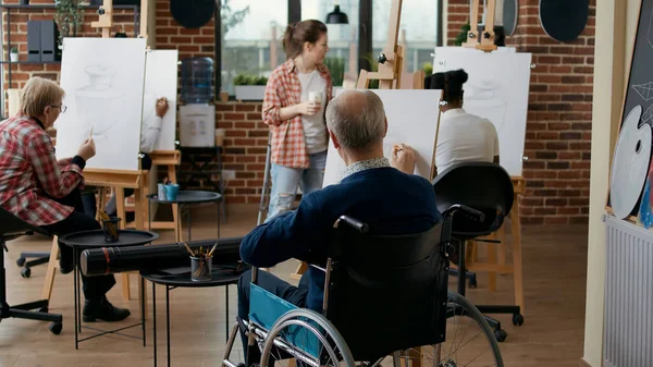 Old man with disability using pencil to draw masterpiece in art class with people. Elder student in wheelchair drawing vase model on canvas, working with artistic tools for personal development.