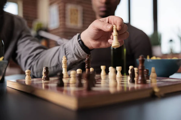 Close up shot of man hand moving chess piece on chessboard while sitting at table. Smart person playing strategy boardgames with friends while sitting at home in living room.