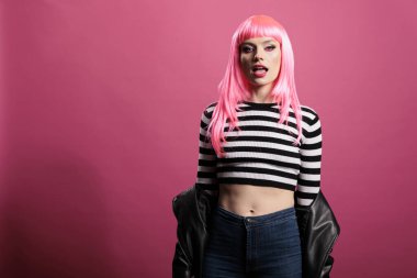 Attractive glamour model with pink hair and leather jacket sitting over studio background, feeling confident and funky with trendy rocker style. Wearing sensual sexy and punk fashion items.