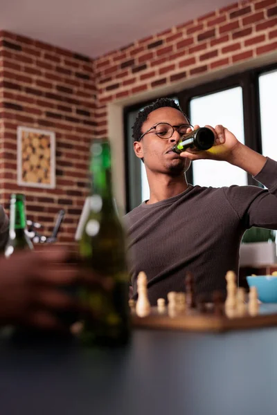 Focused man sipping alcoholic beverage while thinking about next chess move. Intelligent person drinking beer with friends while relaxing at home in living room with chess game and snacks.