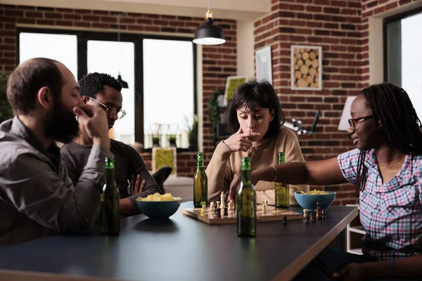 Smart multiethnic group of people sitting at table while playing chess. Happy diverse friends sitting together at home in living room, enjoying strategy boardgames while having beverages and snacks.